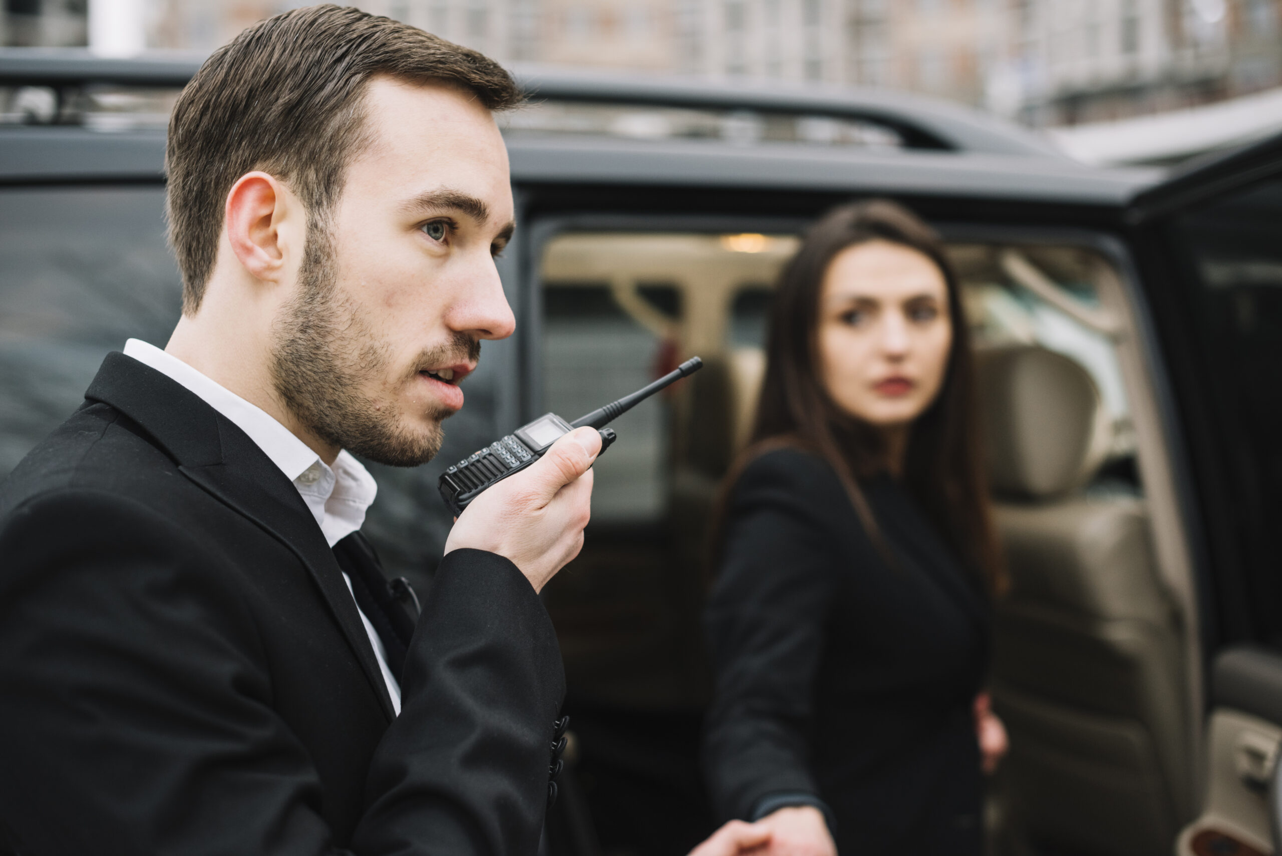 Close protection operative speaking into a radio beside a client vehicle